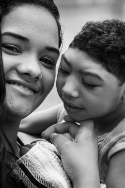 Fotografia em preto e branco de uma jovem mulher que segura no colo uma criança com a síndrome congênita do Vírus Zica. A jovem com o rosto virado para a frente olha para a câmera e sorri, e ela dá apoio ao rosto da criança segurando seu queixo com a mão esquerda.