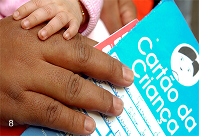 Foto colorida. Detalhe da Mão de uma pessoa de pele escura, que segura um cartão onde está escrito Cartão da Criança. A Mão de um bebê repousa sobre a mão adulta.