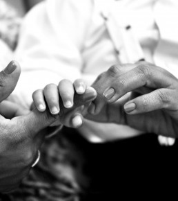 Foto em preto e branco da mão direita de uma criança pequena que segura o dedo indicador da mão direita de uma mulher. A criança está sentada no colo da mulher e está envolvida por seus braços.